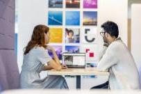 Two employees looking at a laptop seated in a collaboration space, with colourful branded wall pictures in the distance